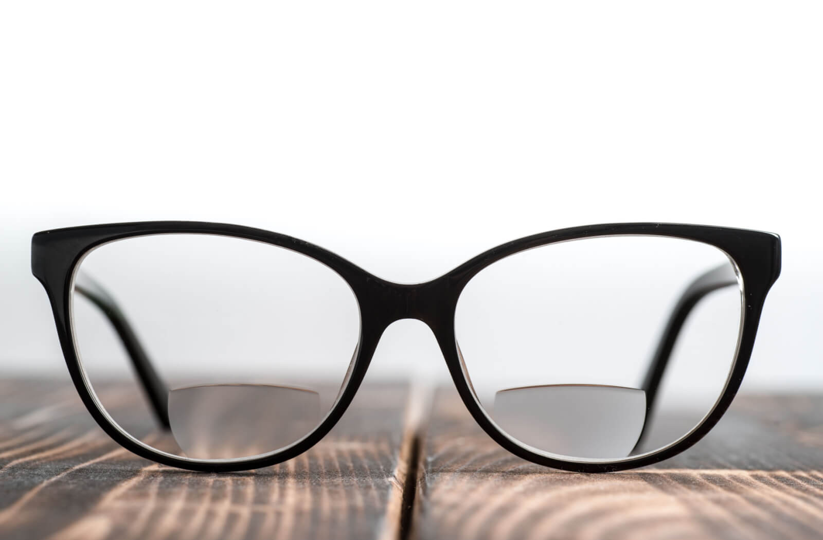 A close-up of black-framed glasses with bifocal lenses sitting on a wooden table