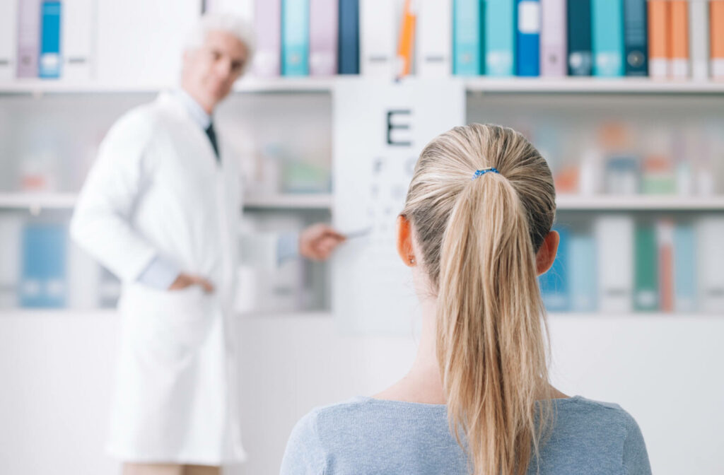 View from behind a woman's head as she sits in a chair and looks ahead while her eye doctor points to an eye chart.