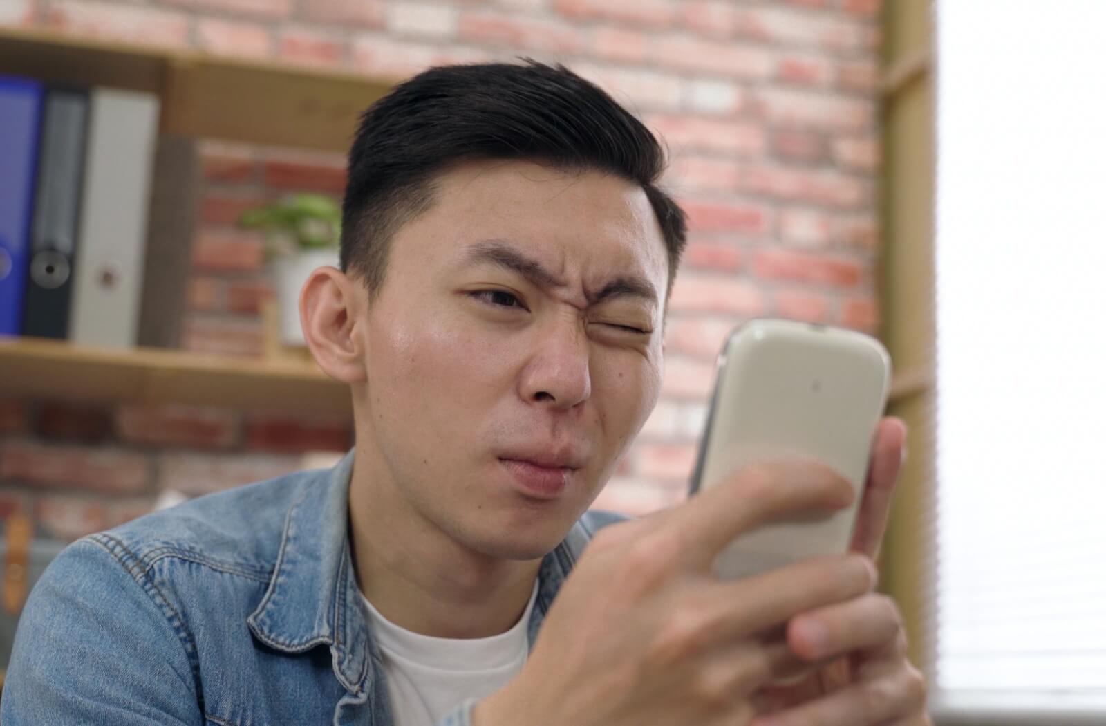 A young adult squints through one eye, holding the other closed to find relief from painful blinking while using their phone
