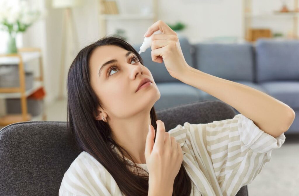 Person sitting indoors and tilting their head back while applying eye drops into one eye.