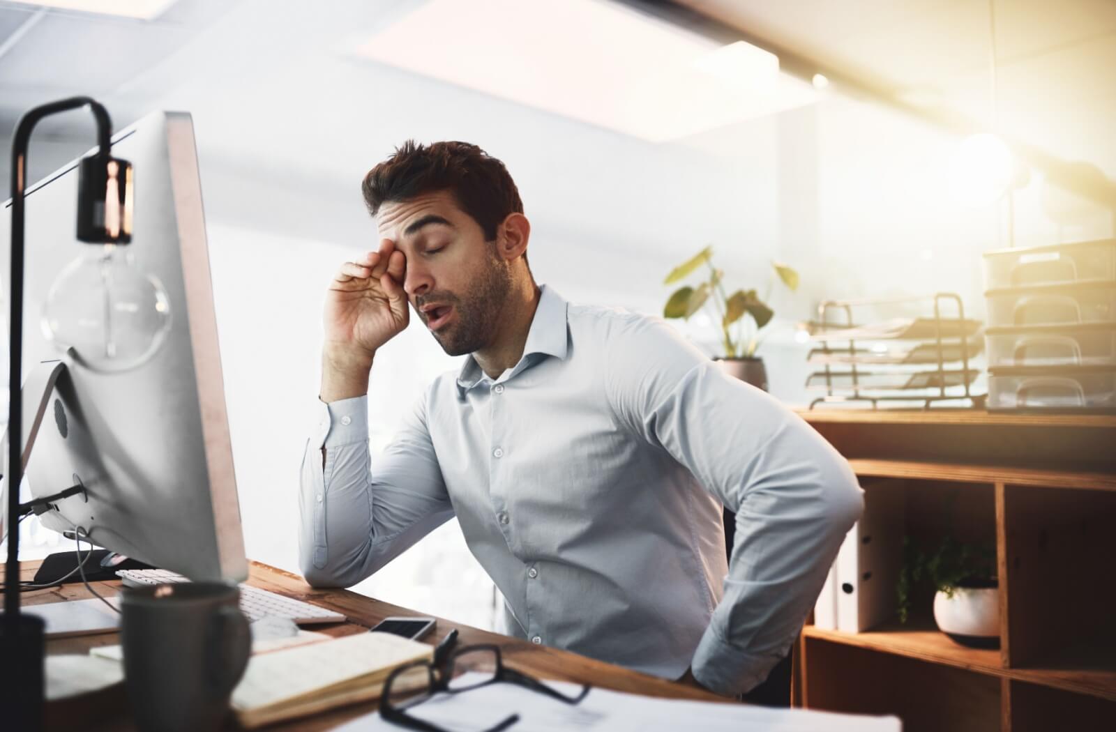 Man sitting at an office desk rubbing his tired eyes while working at a computer, showing signs of eye strain and fatigue.